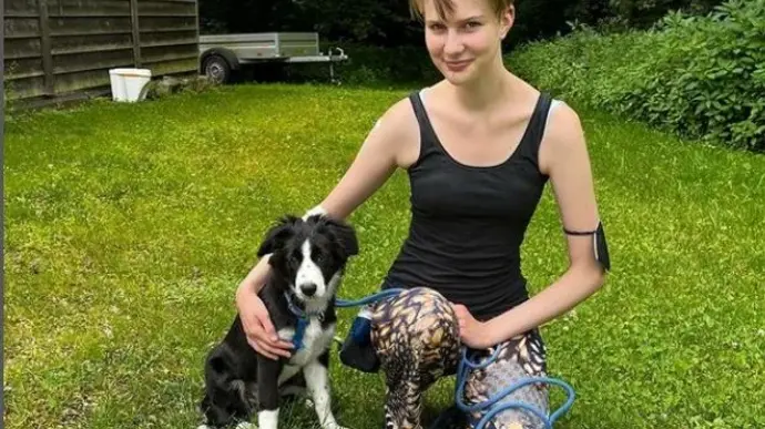 Ein Mädchen sitzt auf dem Rasen und hält einen Hund an der Leine. Sie lächelt und der Hund sitzt neben ihr.