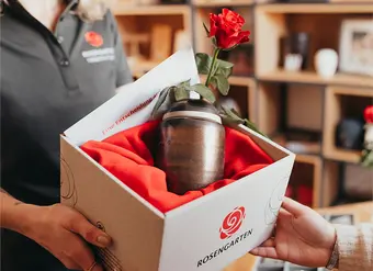 Eine Urne in einer stilvollen Verpackung mit rotem Stoff und Rose, überreicht von einer Rosengarten-Mitarbeiterin