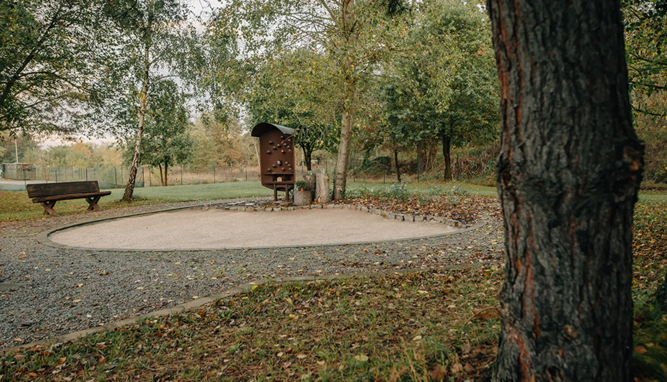 Ein rustikaler Pflanzentrog steht vor einem ruhigen, grünen Rückzugsort mit Gedenkstätte.