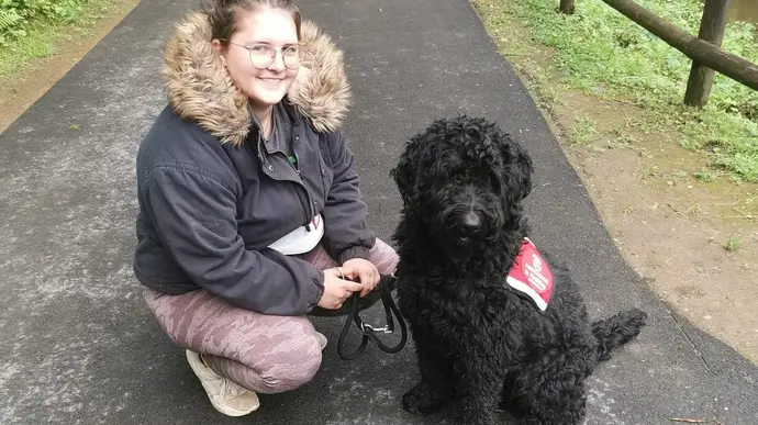 Das Bild zeigt eine junge Frau in einem Park, die auf dem Asphaltweg neben einem schwarzen Hund sitzt.