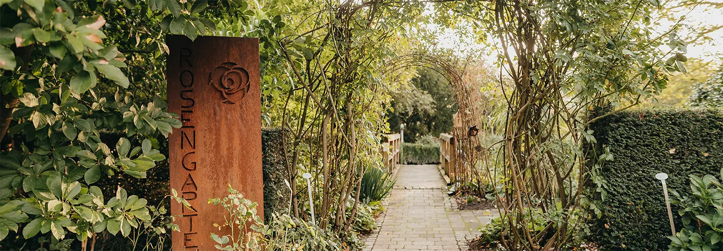 Rostige Metallsäule mit Schriftzug Rosengarten am grünen Eingang eines Gartens