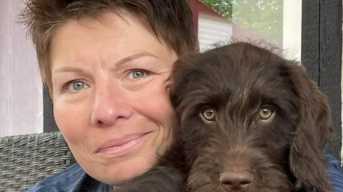 Eine Person hält einen jungen, braunen Hund im Arm. Beide schauen direkt in die Kamera.