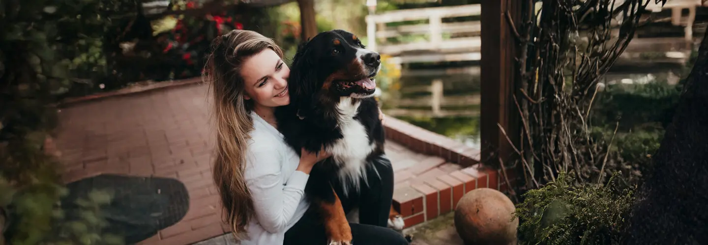 Eine junge Frau schmiegt sich lächelnd an ihren großen Berner Sennenhund auf einer Terrasse.