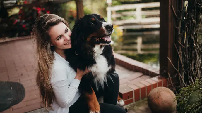 Eine junge Frau schmiegt sich lächelnd an ihren großen Berner Sennenhund auf einer Terrasse.