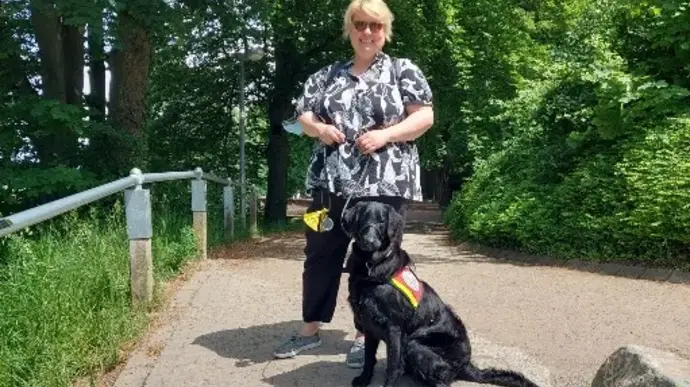 Eine Frau steht auf einem gepflasterten Weg, neben ihr sitzt ein schwarzer Assistenzhund mit Weste.