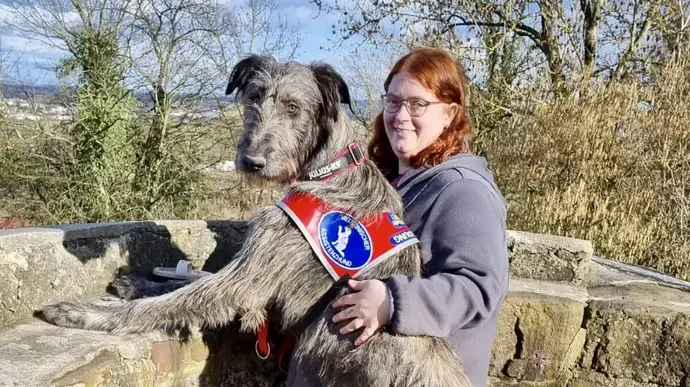 Eine Frau mit ihrem großen Wolfshund, der mit den Vorderpfoten auf eine Mauer lehnt