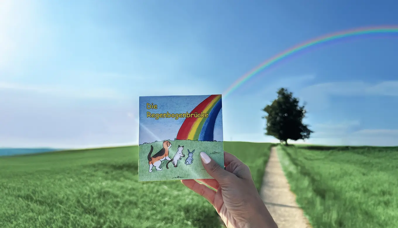 Ein liebevoll gestaltetes Kinderbüchlein mit dem Titel „Die Regenbogenbrücke“, das Tiere auf ihrem Weg über einen bunten Regenbogen zeigt und Trost sowie Hoffnung vermittelt, gehalten vor einer grünen Wiesenlandschaft mit echtem Regenbogen am Himmel – im ROSENGARTEN-Tierkrematorium