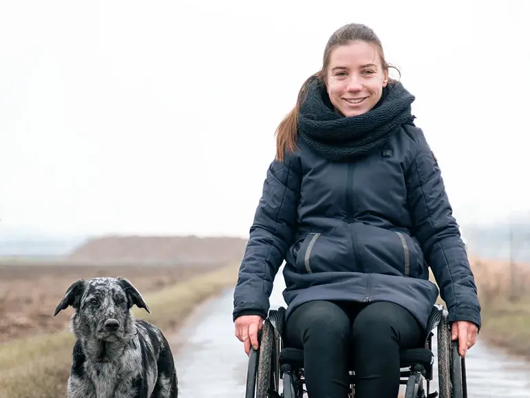 Eine Person im Rollstuhl fährt auf einem nassen Feldweg, begleitet von einem Hund, der neben ihr läuft. 