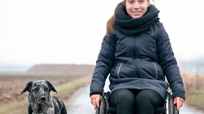 Eine Person im Rollstuhl fährt auf einem nassen Feldweg, begleitet von einem Hund, der neben ihr läuft. 