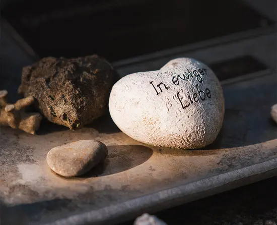 Weißer Gedenkstein mit der Inschrift „In ewiger Liebe“, umgeben von kleinen Natursteinen auf einer Holzfläche