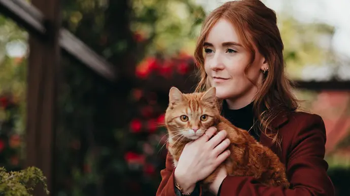 Eine junge Frau hält eine rote Katze im Arm und blickt ruhig in die Ferne.
