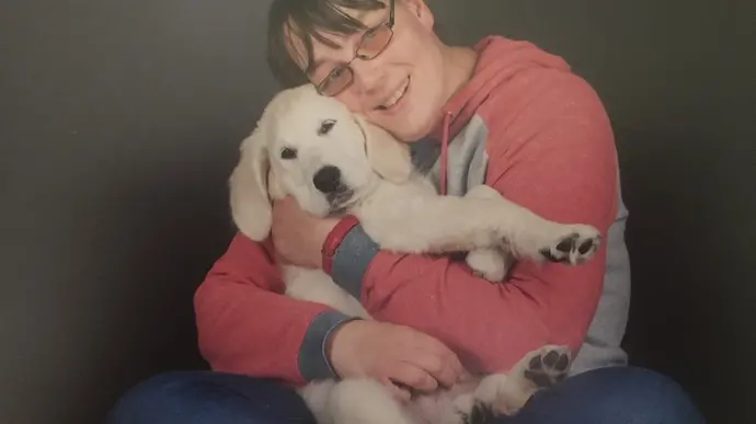 Eine Person umarmt einen Labrador-Welpen, während sie auf dem Boden sitzt. Beide blicken zur Kamera.